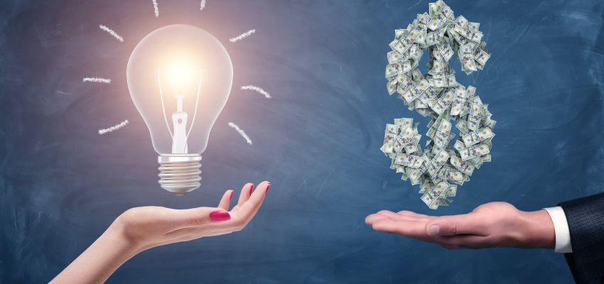 a hand holds a glowing light bulb, next to another hand holding money