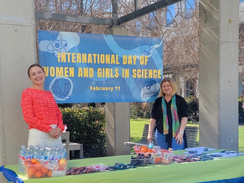 Two woman smile in front of a banner for woman in science day at uci